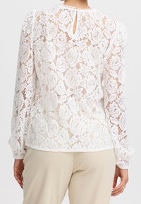 Blusa bianca in pizzo con motivo floreale, scollo rotondo e dettaglio a chiave sulla schiena. Maniche lunghe con polsini arricciati. Indossata sopra pantaloni beige.