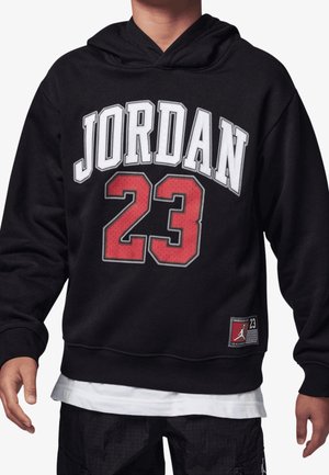 Czarna bluza z kapturem z białym napisem "JORDAN" i dużą czerwoną liczbą "23", noszona na białej koszulce i czarnych spodniach, widok z przodu na tors i ręce.