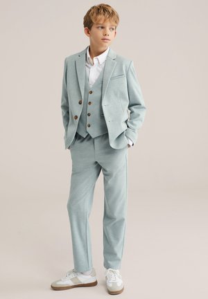 Svijetloplavo odijelo od mekane tkanine, s dvostrukim prslukom, krojenim sako i hlačama u istoj boji, u kombinaciji s bijelim tenisicama.