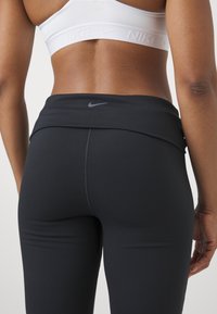 Gros plan du bas du dos d'une personne portant un soutien-gorge de sport Nike blanc et des leggings noirs taille haute Nike avec logos visibles.