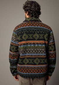 Gehaakte fleece jas met een relaxte pasvorm, voorzien van multicolore geometrische patronen in groen, oranje en blauw op een zwarte achtergrond.