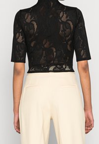 Schwarzes Spitzencrop-Top mit kurzen Ärmeln, hohem Kragen und transparenten floralem Mustern. Kombiniert mit hellbeigen, maßgeschneiderten Hosen.
