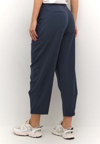 Pantalon bleu marine à coupe ajustée, doté d'une poche arrière et d'évents latéraux, porté avec des baskets blanches et grises.