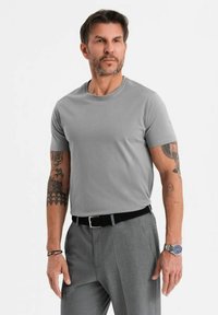 Grå bomullströja med rund halsringning, matchad med grå byxor. Har tatueringar på armarna och ett silverur med blå urtavla.