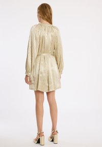 Robe à manches courtes, plissée, en tissu métallisé doré avec des rayures texturées et une ceinture à la taille. Des sandales à talons épais complètent le look.