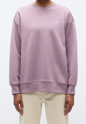 Personne portant un sweat-shirt à manches longues violet clair avec un petit logo circulaire blanc sur la poitrine et un pantalon beige.