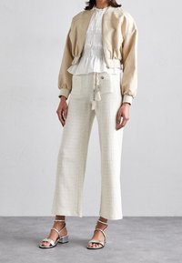 Vrouw in een beige gek cropped jack, witte gestructureerde wijde broek met een kwastceintuur, en zilveren open teen enkelband sandalen die op een grijze vloer staat.