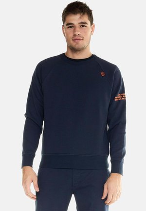 Junger Mann, der einen marineblauen Sweatshirt mit Rundhalsausschnitt und passende Hose trägt, steht vor einem schlichten hellen Hintergrund.