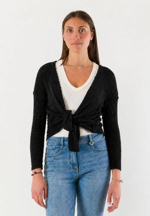 Donna con lunghi capelli castani che indossa una maglietta bianca a V, un cardigan nero texturizzato legato in vita e jeans blu, in piedi davanti a uno sfondo semplice.