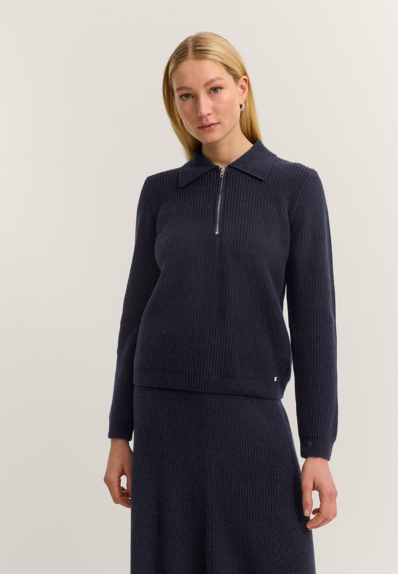 Marineblaues, geripptes Strickpullover mit Reißverschlusskragen, langen Ärmeln und geradem Schnitt. Teil eines koordinierten Outfits mit einem passenden Rock.