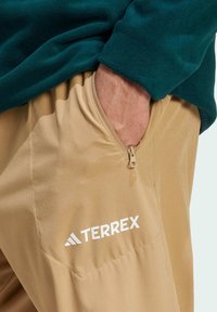 Beige Terrex-Hose mit einer schrägen Reißverschlusstasche und weißen Logodetails. Weiche Textur, kombiniert mit einem dunkelblauen Fleece-Oberteil, das ein geschichtetes Design präsentiert.
