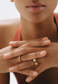 Anillos de oro con acentos esféricos en varios dedos, reposando sobre una piel ligeramente texturizada. Un esmalte de uñas neutro realza una estética minimalista.