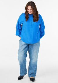 Usmívající se žena s dlouhými hnědými vlasy má na sobě zářivě modrou pruhovanou blůzu, volné světle modré džíny a černé boty, má ruce v kapsách a dívá se dolů.