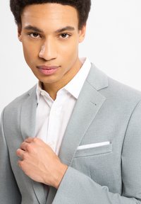 Hellgrauer Blazer aus strukturiertem Stoff, mit einer einzelnen Brusttasche mit weißem Rand, kombiniert mit einem weißen Hemd. Nahaufnahme.