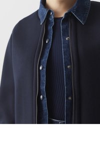 Mörkblå zip-up jacka med en denimkrage. Har tryckknappar, en slät textur och en ribbad mörkblå topp under.