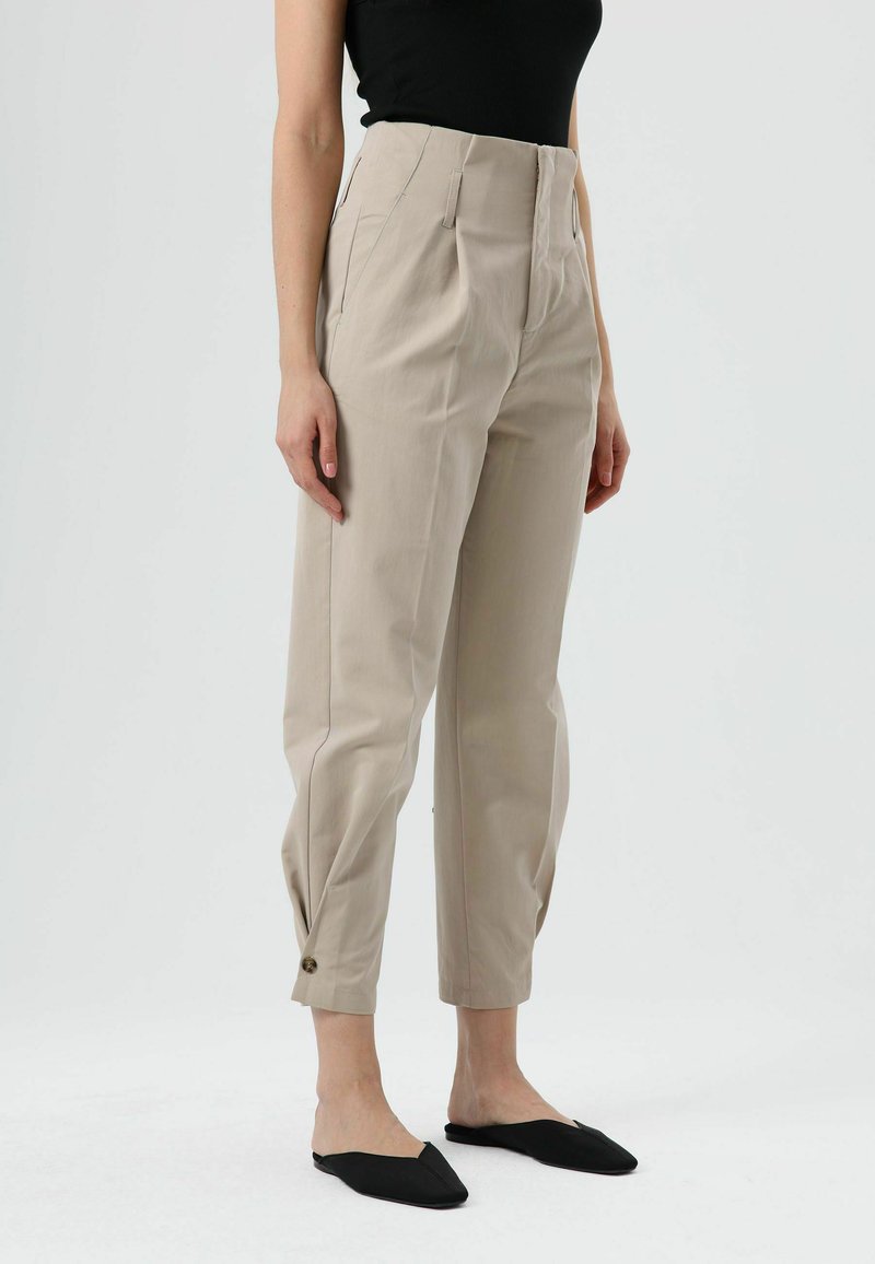 Pantalon beige taille haute avec une coupe ajustée, poches latérales et détails à boutons à l'ourlet, associé à des ballerines noires sans lacets.