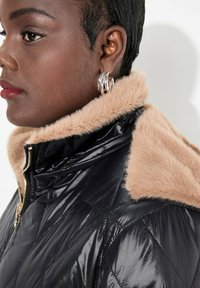 Zwarte gewatteerde pufferjack met een tan faux fur kraag, voorzien van een ritssluiting en zilveren hardware accenten. Glad, glanzend textuur.