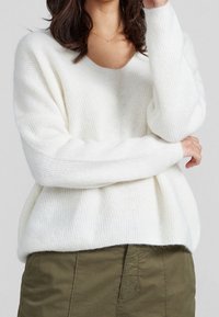 Pull blanc en maille avec col en V et coupe ample, présentant une texture côtelée et des manches larges, associé à une jupe vert olive.