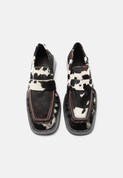 Pár čiernych a bielych mokasín s vzorom hovädzej kože, štvorcovými špičkami a hrubými čiernymi podrážkami na jednofarebnom pozadí.