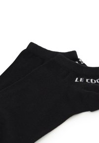 Calcetines negros hasta el tobillo, fabricados en un tejido suave con puño acanalado; presentan el logotipo en texto blanco "LE COQ SPORTIF" cerca de la abertura.