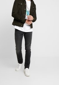 Chaqueta de pana verde oscuro con detalles de botones, combinada con una camiseta gráfica blanca y jeans negros ajustados, complementados con zapatillas blancas.