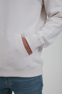 Hellgrauer Kapuzenpullover mit Kängurutasche, gerippten Bündchen und Taillenbund; kombiniert mit blauen Jeans. Die weiche Stoffstruktur ist sichtbar.