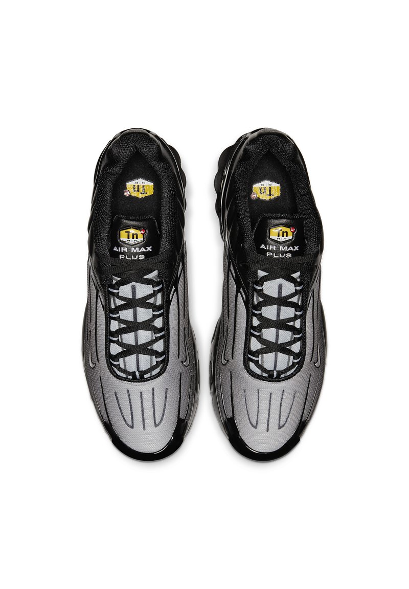 Zapatillas deportivas negras y grises con una parte superior de malla, diseño Curvo y un sistema de cordones pronunciado. Presenta la marca "Air Max Plus".