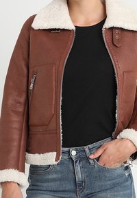 Giubbotto in pelle marrone con colletto in shearling bianco, chiusura con zip frontale e tasche laterali. Finitura testurizzata con cuciture a contrasto e polsini.