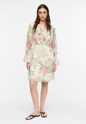 Kobieta ubrana w kwiatową sukienkę do kolan z długimi przezroczystymi rękawami, falbanami i czarnymi płaskimi sandałami, stojąca na jednolitym białym tle.