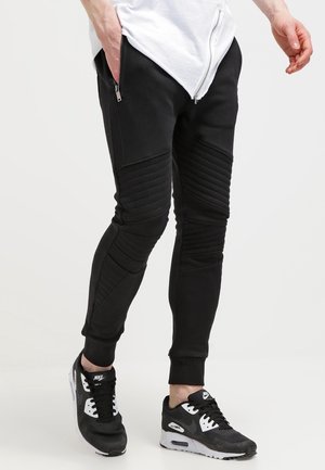 Schwarze, taillierte Jogginghosen aus weichem Material, mit gerippten Knieeinsätzen und seitlichen Reißverschlusstaschen. Kombiniert mit schwarz-weißen Sportschuhen.