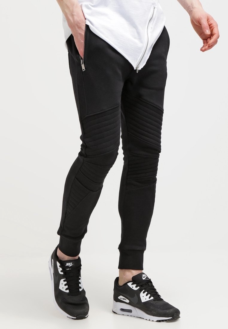 Černé tapered tepláky vyrobené z jemného materiálu, s vyztuženými kolenními detaily a bočními zipovými kapsami. V kombinaci s černobílými sportovními teniskami.