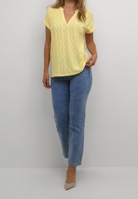 Keltainen lyhythihainen toppi, jossa on V-pääntie ja aaltoileva rei'itetty kuvio, yhdistettynä vaaleansinisiiin korkeavyötäröisiin farkkuihin ja beigeihin korkokenkiin.