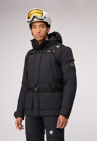 Zwarte geïsoleerde winterjas met een capuchon, verstelbare riem en meerdere zakken. Gecombineerd met zwarte broek met een logo op het been.