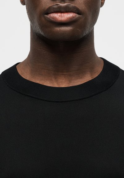 Gros plan sur le bas du visage et le cou d'une personne à la peau foncée portant un t-shirt noir uni à col rond, sur un fond blanc.