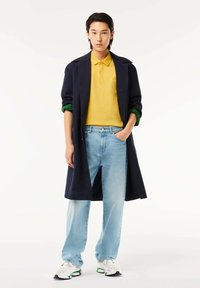 Marinblå lång kappa, gul polo tröja, ljusblå jeans och vita sneakers. Kappan har en skräddarsydd design med gröna manschetter.