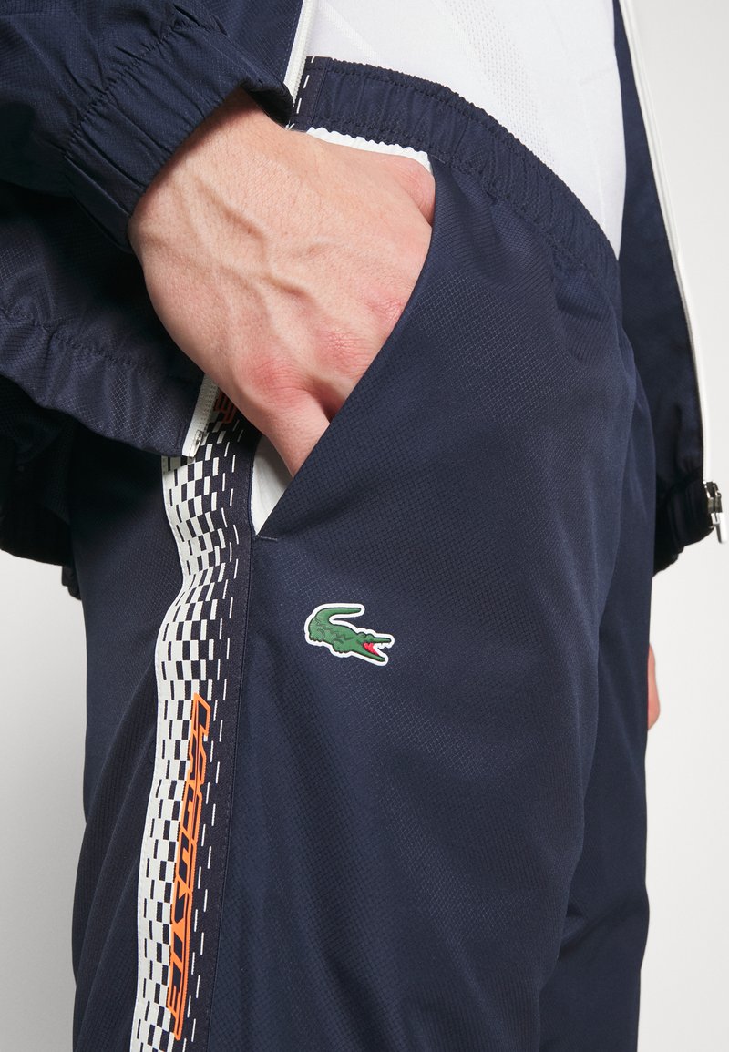 Pantalons de sport navy en tissu texturé, avec un design à carreaux blancs sur le côté et un logo de crocodile vert sur la poche.