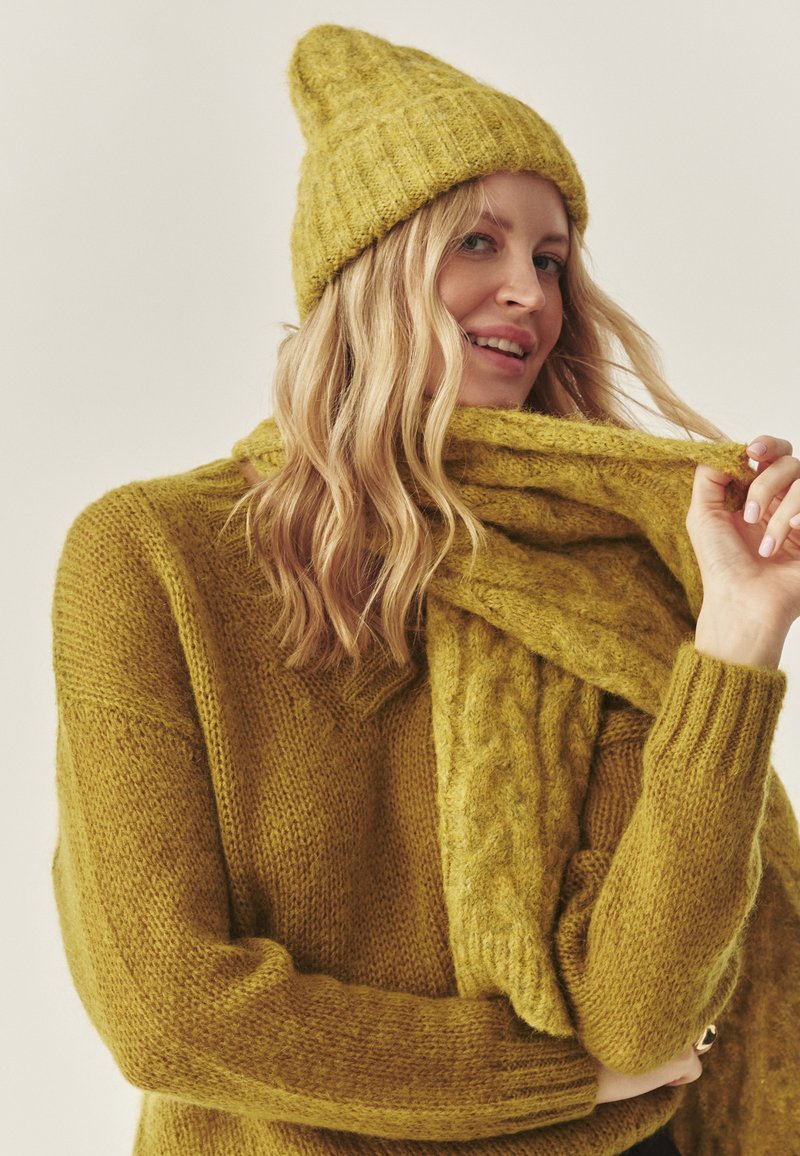 Maglione giallo senape lavorato a maglia con cappellino abbinato, entrambi con dettagli a coste. Una sciarpa testurizzata avvolge il collo, aggiungendo calore.