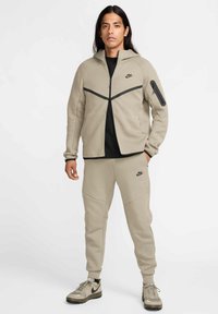Beige Nike-Jacke mit schwarzen Akzenten und vollwertigem Reißverschluss; kombiniert mit passenden beigen Hosen mit geschwungenen Nahtdetails. Strukturierter Stoff.