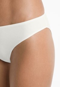 Fehér bordás bikini alsó alacsony szabású dizájnnal. Sima textúra, kivágott lábnyílások és minimalista takarás. Ideális fürdőruhának vagy strandszóknak.
