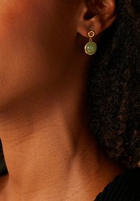 Gouden oorbellen met een ronde groene edelsteen, hangend aan een ronde studs. Het ontwerp benadrukt gladde texturen en een minimalistische stijl.