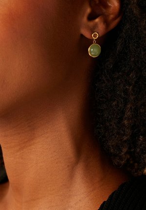 Gouden oorbellen met een ronde groene edelsteen, hangend aan een ronde studs. Het ontwerp benadrukt gladde texturen en een minimalistische stijl.