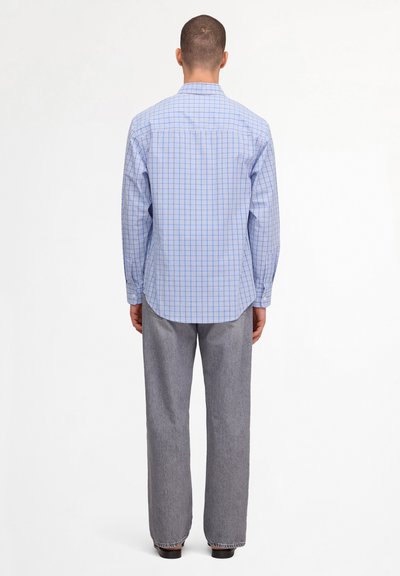 Homme portant une chemise bleue à carreaux manches longues et un pantalon gris ample, debout de dos sur un fond blanc uni.
