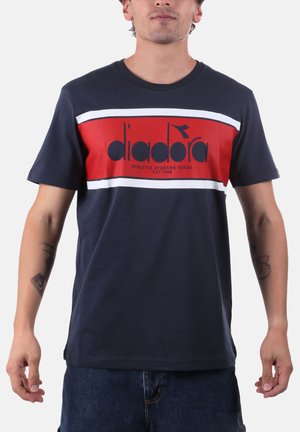 Uomo con t-shirt blu navy con banda orizzontale rossa e bianca e logo "diadora", abbinata a pantaloncini di jeans blu scuro.