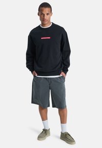 Zwarte sweatshirt met een rood Quiksilver-logo, gecombineerd met grijze shorts en lichtgrijze schoenen. Model staat met handen in de zakken.