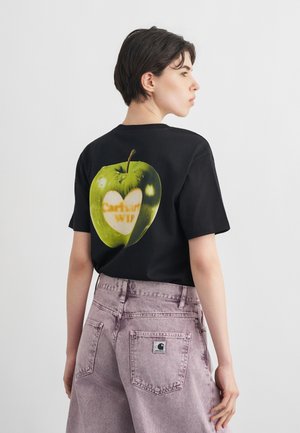 Žmogus su trumpais tamsiais plaukais dėvi juodą marškinėlį su žaliu obuoliu, kuriame yra baltos spalvos širdis, ir dalinai matomu tekstu gale.