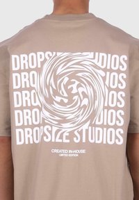 Smeđi pamučni majica s grafikom spirale i ponovljenim natpisom "DROPSIZE STUDIOS" u bijeloj boji, s natpisom "STVORENO U KUĆI OGRANIČENA EDICIJA" ispod.