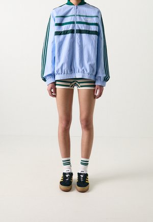 Persoon met lichtblauwe jas met ritssluiting, groene en beige gestreepte korte broek, witte sokken met groene strepen en zwarte sneakers met gele strepen.