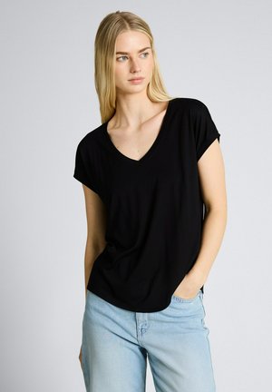 Blondine draagt een zwarte V-hals top met korte mouwen en lichtblauwe jeans, staat met één hand in de zak en kijkt naar links.