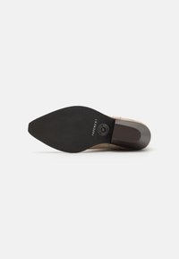 Pantof bej cu toc înalt, vârf ascuțit și superior din suede texturat. Talpa din cauciuc negru are o ușoară curbură și un branding embosat.
