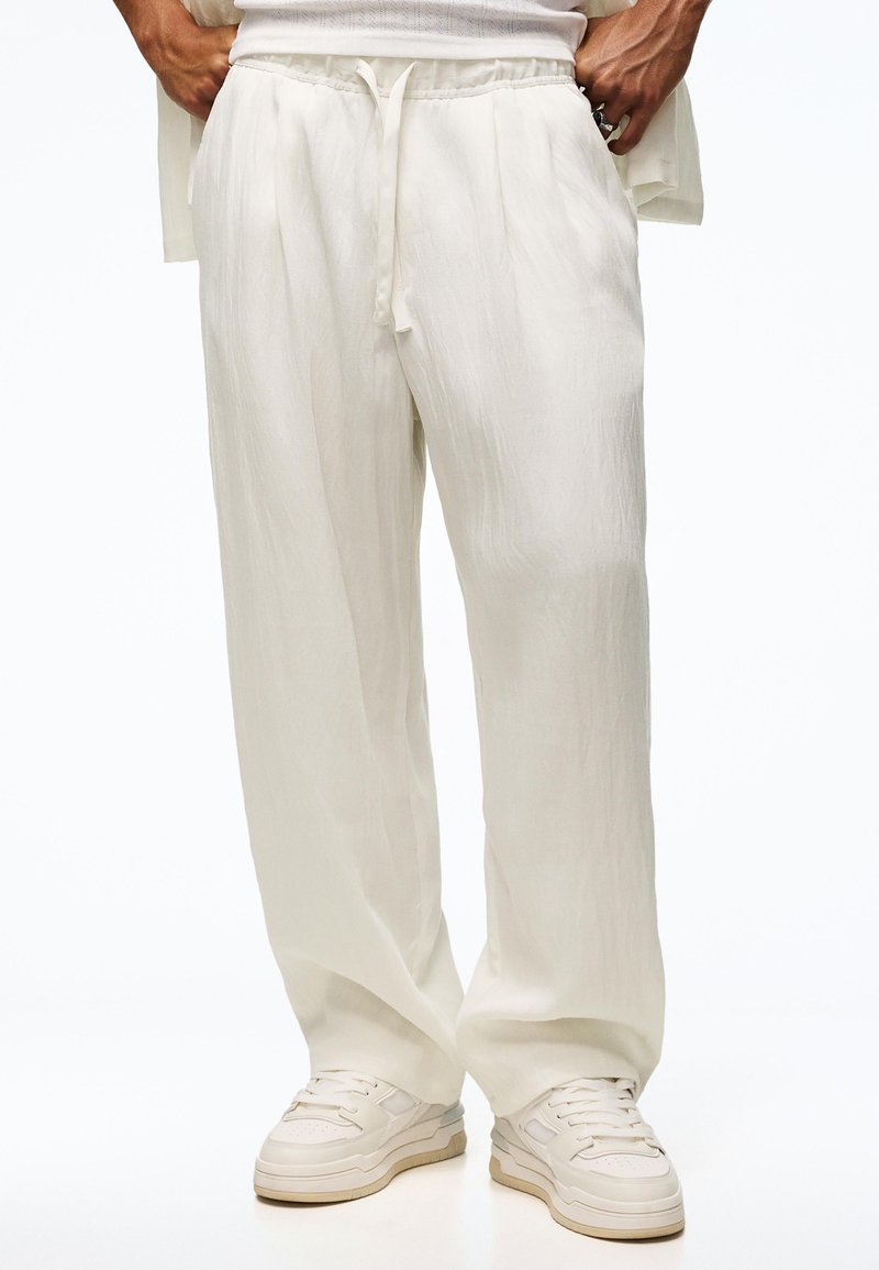 Personne portant un pantalon ample écru à cordon et des baskets blanches, debout les mains dans les poches sur un fond uni.